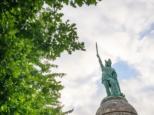 Hermannsdenkmal in Detmold vor bewölktem Himmel, von Bäumen eingerahmt, erhobenes Schwert.