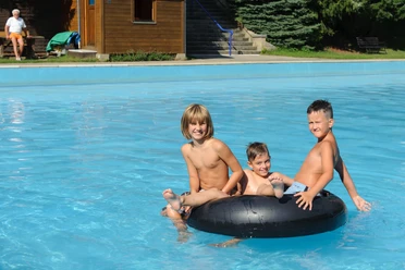 Drie kinderen zitten te lachen op een zwarte zwemband in een blauw zwembad; een oudere man zit op een bankje op de achtergrond.