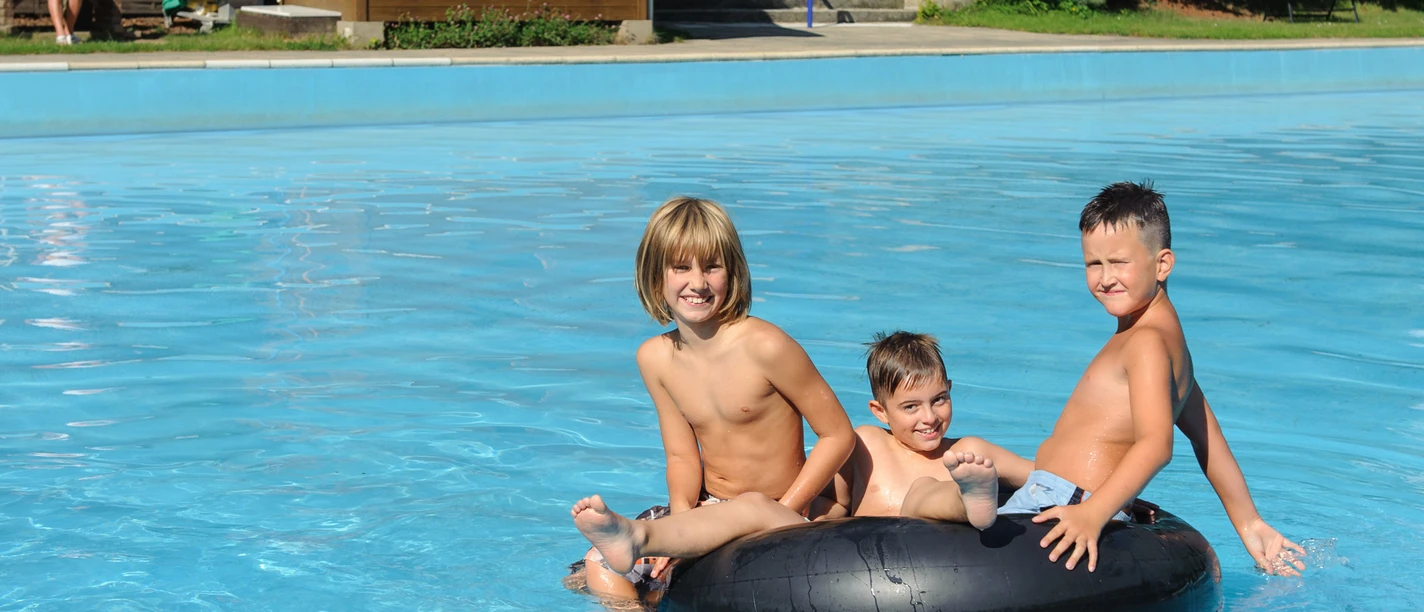 Drie kinderen zitten te lachen op een zwarte zwemband in een blauw zwembad; een oudere man zit op een bankje op de achtergrond.