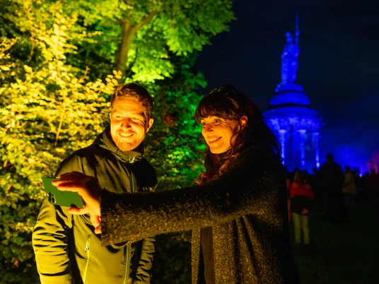 Detmold-Der Hermann leuchtet-Teutoburger-Wald-Tourismus-D-Ketz-105.jpg Zwei Personen machen ein Selfie vor dem abends angestrahlten Hermannsdenkmal in einem Park.