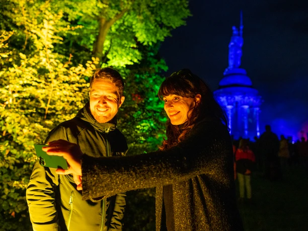 Detmold-Der Hermann leuchtet-Teutoburger-Wald-Tourismus-D-Ketz-105.jpg Zwei Personen machen ein Selfie vor dem abends angestrahlten Hermannsdenkmal in einem Park.