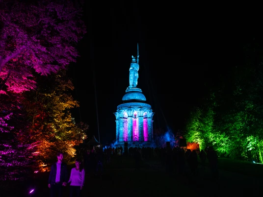 Detmold-Der Hermann leuchtet-Teutoburger-Wald-Tourismus-D-Ketz-112.jpg Das beleuchtete Hermannsdenkmal in Detmold in bunten Farben bei Nacht; Menschenmengen im Vordergrund.