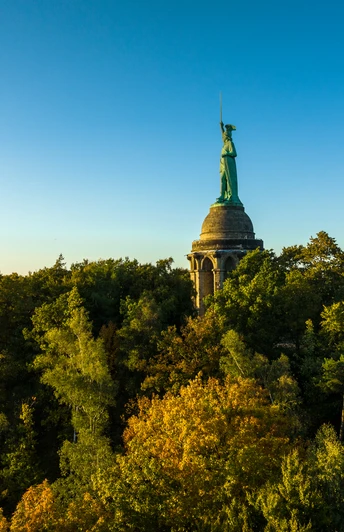 Denkmal auf bewaldetem Hügel bei Sonnenuntergang, im Hintergrund blauer Himmel.