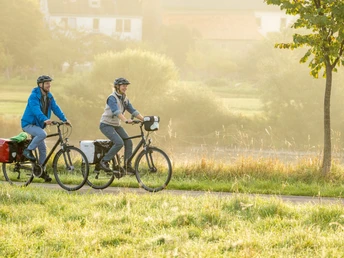 Zwei Personen radeln auf einem Pfad durch eine ländliche, sonnenbeschienene Landschaft.