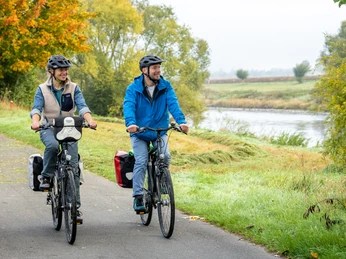 Zwei Radfahrer auf einem asphaltierten Weg entlang eines herbstlichen Flussufers in ländlicher Idylle.