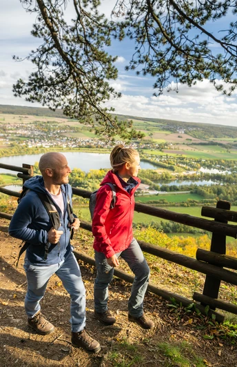 Zwei Wanderer mit Rucksack genießen einen Panoramablick auf eine Flusslandschaft bei sonnigem Wetter.