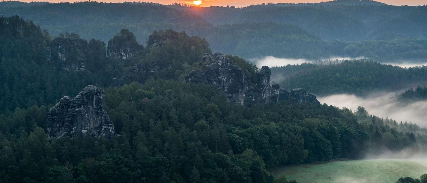 Sonnenaufgang im Basteigebiet Sonnenaufgang über einer nebligen Waldlandschaft mit markanten Felsformationen im Vordergrund.