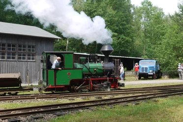 Green steam locomotive with smoke runs on rails in front of a gray building, a blue truck stands next to it; people watch the scene.
