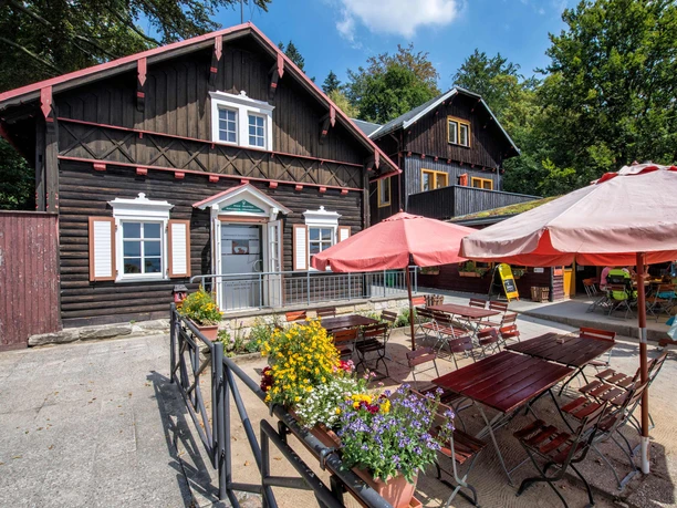 A rustic wooden house with red accents, surrounded by trees, with a cozy outdoor area with tables, chairs and colorful flowers under umbrellas.