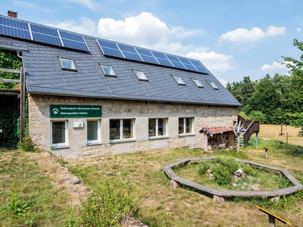 Stenen huis met zonnepanelen op het dak, omgeven door groene weide en bomen, met een bord "Nationalpark Sächsische Schweiz Bildungsstätte Sellnitz".