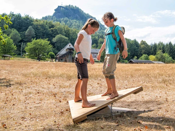 Twee kinderen balanceren op blote voeten op een houten plank in de buitenlucht, omringd door een weiland, bomen en een huis op de achtergrond.