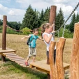 Bildungsstätte Sellnitz Zwei Kinder balancieren auf einem hölzernen Kletterparcours im Freien, umgeben von Bäumen und einer Wiese unter blauem Himmel.Two children balance on a wooden climbing course outdoors, surrounded by trees and a meadow under a blue sky.Dvě děti balancují na dřevěné prolézačce venku, obklopené stromy a loukou pod modrou oblohou.Dwoje dzieci balansuje na drewnianym torze wspinaczkowym na zewnątrz, otoczonym drzewami i łąką pod błękitnym niebem.Twee kinderen balanceren op een houten klimparcours buiten, omringd door bomen en een weiland onder een blauwe hemel.Due bambini in equilibrio su un percorso di arrampicata in legno all'aperto, circondati da alberi e da un prato sotto un cielo azzurro.