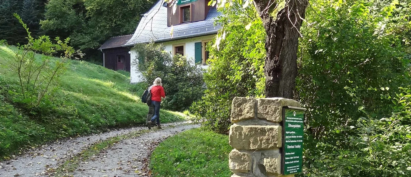 Infostelle Zeughaus Eine Person in roter Jacke geht einen Weg entlang zu einem weißen Haus mit grünem Dach, umgeben von üppiger Vegetation und Bäumen.