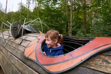Waldhusche Hinterhermsdorf Ein lachendes Kind in blauer Jacke schaut aus einem hölzernen Spielgerät, das wie ein Insekt gestaltet ist, in einem Wald.