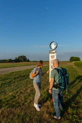 Zwei Wanderer lesen ein Schild auf einem ländlichen Weg bei Sonnenschein, blauer Himmel im Hintergrund.