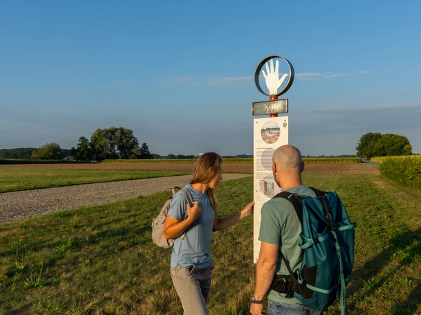 Delbrueck-Roemerlager-Teutoburger-Wald-Tourismus-Patrick-Gawandtka-017.jpg Zwei Wanderer lesen ein Schild auf einem ländlichen Weg bei Sonnenschein, blauer Himmel im Hintergrund.