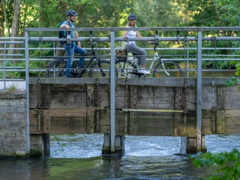 Zwei Radfahrer auf einer Brücke im grünen Park, darunter fließt ein kleiner Fluss.