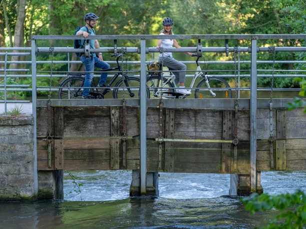 Delbrueck-BokerKanal-Teutoburger-Wald-Tourismus-Patrick-Gawandtka-036.jpg Zwei Radfahrer auf einer Brücke im grünen Park, darunter fließt ein kleiner Fluss.