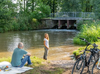 Eine Frau steht am Ufer eines Flusses neben einem Mann auf einer Decke, Fahrräder sind abgestellt.