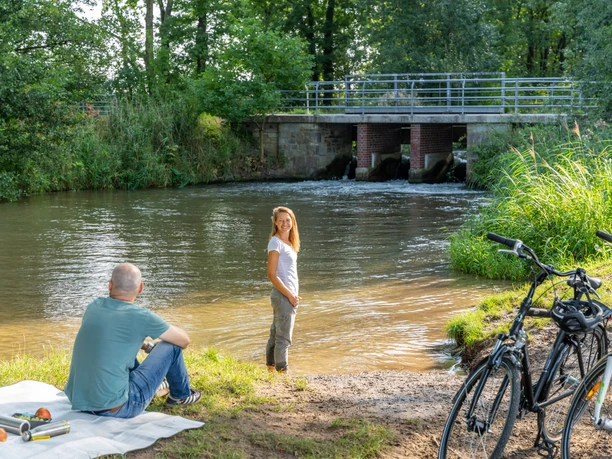 Eine Frau steht am Ufer eines Flusses neben einem Mann auf einer Decke, Fahrräder sind abgestellt.