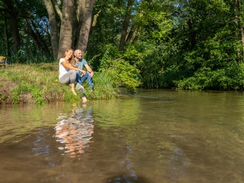 Delbrueck-BokerKanal-Teutoburger-Wald-Tourismus-Patrick-Gawandtka-047.jpg Paar sitzt am Flussufer in einem sonnendurchfluteten Wald, lächelnd und entspannend im Grünen.