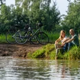 Ein Paar sitzt entspannt am Flussufer, im Hintergrund stehen Fahrräder, umgeben von grüner Natur.