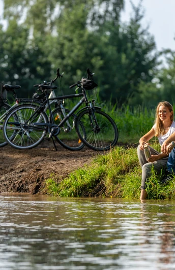 Ein Paar sitzt entspannt am Flussufer, im Hintergrund stehen Fahrräder, umgeben von grüner Natur.