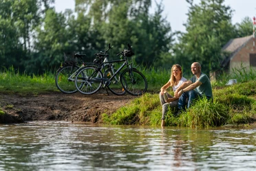 Delbrueck-BokerKanal-Teutoburger-Wald-Tourismus-Patrick-Gawandtka-049.jpg Ein Paar sitzt entspannt am Flussufer, im Hintergrund stehen Fahrräder, umgeben von grüner Natur.