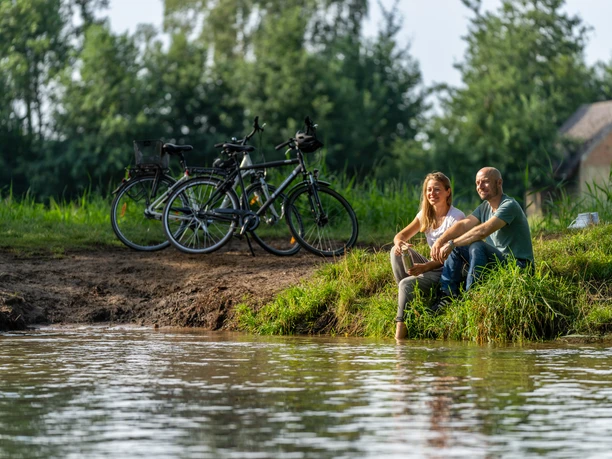 Delbrueck-BokerKanal-Teutoburger-Wald-Tourismus-Patrick-Gawandtka-049.jpg Ein Paar sitzt entspannt am Flussufer, im Hintergrund stehen Fahrräder, umgeben von grüner Natur.