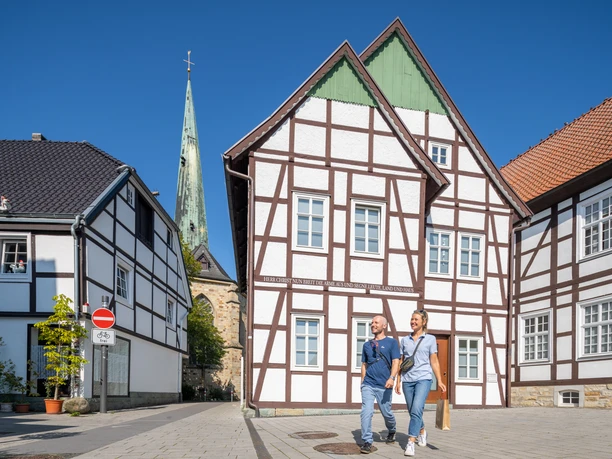 Zwei Personen spazieren fröhlich vor historischen Fachwerkhäusern, im Hintergrund ragt ein Kirchturm.