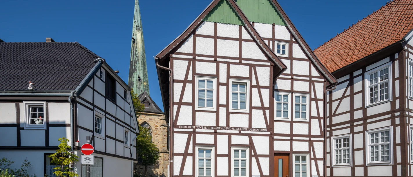 Delbrueck-Innenstadt-Teutoburger-Wald-Tourismus-Patrick-Gawandtka-056-CC-BY-SA.jpg Fachwerkhäuser vor blauem Himmel mit Kirchturm im Hintergrund, gepflasterte Straße ohne Verkehr.
