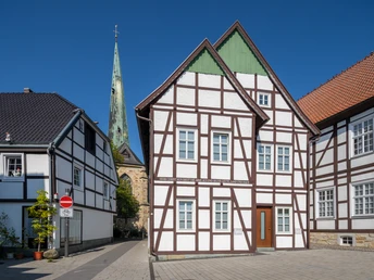 Delbrueck-Innenstadt-Teutoburger-Wald-Tourismus-Patrick-Gawandtka-056-CC-BY-SA.jpg Fachwerkhäuser vor blauem Himmel mit Kirchturm im Hintergrund, gepflasterte Straße ohne Verkehr.