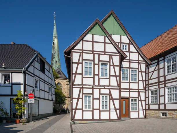 Delbrueck-Innenstadt-Teutoburger-Wald-Tourismus-Patrick-Gawandtka-056-CC-BY-SA.jpg Fachwerkhäuser vor blauem Himmel mit Kirchturm im Hintergrund, gepflasterte Straße ohne Verkehr.