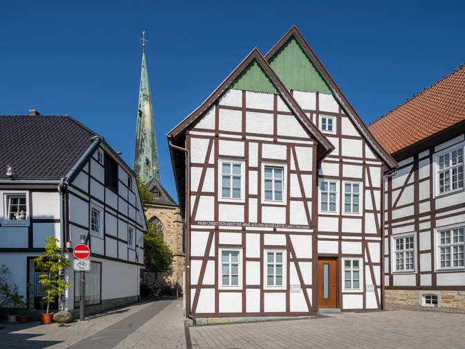 Delbrueck-Innenstadt-Teutoburger-Wald-Tourismus-Patrick-Gawandtka-056-CC-BY-SA.jpg Fachwerkhäuser vor blauem Himmel mit Kirchturm im Hintergrund, gepflasterte Straße ohne Verkehr.