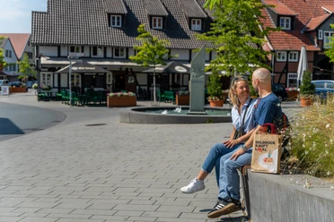Ein Paar sitzt lachend auf einer Parkbank vor traditionellen Fachwerkhäusern in einer Fußgängerzone.