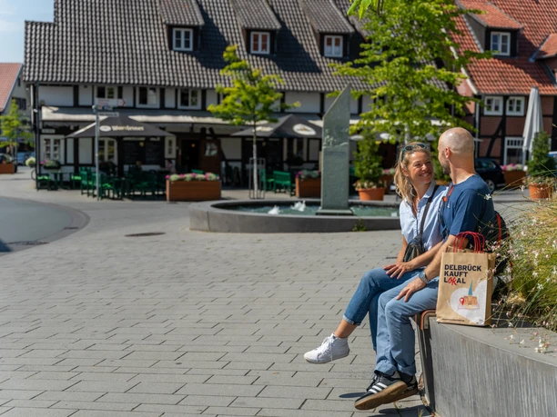 Ein Paar sitzt lachend auf einer Parkbank vor traditionellen Fachwerkhäusern in einer Fußgängerzone.