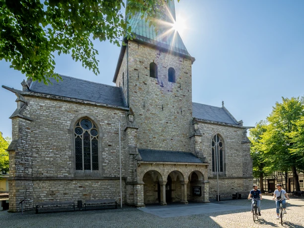 Fahrradfahrer vor historischer Kirche mit hohem Turm unter klarem, sonnigem Himmel, umgeben von Bäumen.