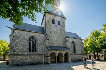 Delbrueck-Kirchplatz-Teutoburger-Wald-Tourismus-Patrick-Gawandtka-060.jpg Fahrradfahrer vor historischer Kirche mit hohem Turm unter klarem, sonnigem Himmel, umgeben von Bäumen.