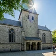 Delbrueck-Kirchplatz-Teutoburger-Wald-Tourismus-Patrick-Gawandtka-060.jpg Fahrradfahrer vor historischer Kirche mit hohem Turm unter klarem, sonnigem Himmel, umgeben von Bäumen.