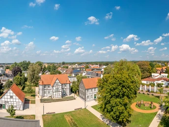 Dorfpanorama mit Fachwerkhäusern, Bäumen und modernen Gebäuden unter blauem Himmel mit Wolken.