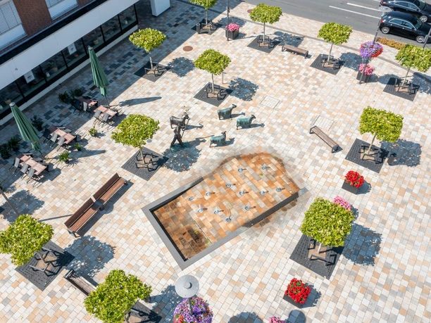 Ein Platz mit gepflasterten Boden, Schachbrettmuster, Brunnen in der Mitte, Bäume und Sitzgelegenheiten.