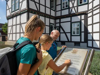 Drei Personen lesen eine Infotafel vor einem Fachwerkhaus an einem sonnigen Tag.