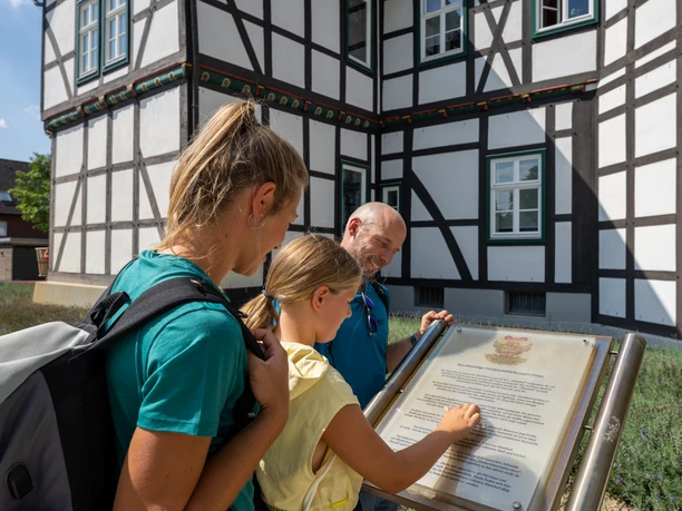 Hoevelhof-Schlossgarten-Teutoburger-Wald-Tourismus-Patrick-Gawandtka-084.jpg Drei Personen lesen eine Infotafel vor einem Fachwerkhaus an einem sonnigen Tag.