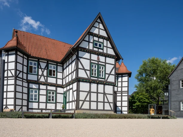 Fachwerkhaus mit rotem Ziegeldach und grünen Fenstern vor klarem, blauem Himmel, umgeben von Bäumen.