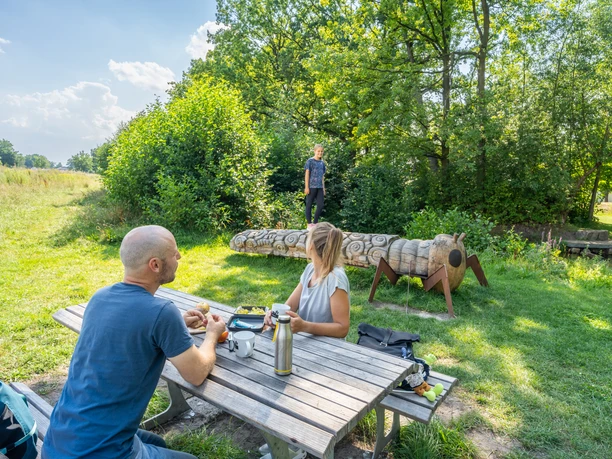 Hoevelhof-Krollbach-Bifurkation-Teutoburger-Wald-Tourismus-Patrick-Gawandtka-106.jpg Ein Paar sitzt an einem Picknicktisch im Grünen, während ein Kind auf einer Baumstamm-Skulptur balanciert.