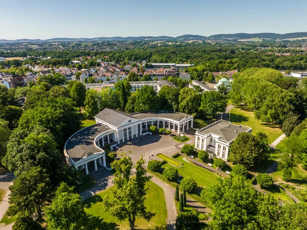 Bad Oeynhausen-Kurpark-Teutoburger-Wald-Tourismus-D-Ketz-084-CC-BY-SA.jpg Kurhaus Bad Oeynhausen: Historischer Bau mit Rundbogenfassade, umgeben von grüner Parklandschaft.