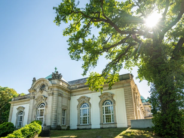 Bad Oeynhausen-Kurpark-Teutoburger-Wald-Tourismus-D-Ketz-076-CC-BY-SA.jpg Historisches Gebäude mit verzierten Fenstern, umgeben von grünem Garten und strahlendem Sonnenlicht.
