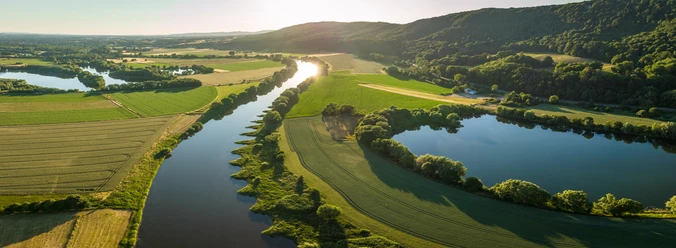 Porta Westfalica-Weserbogen-Teutoburger-Wald-Tourismus-D-Ketz-048.jpg Luftaufnahme eines malerischen Flusslaufs inmitten grüner Felder und bewaldeter Hügel bei Sonnenuntergang.