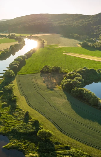 Porta Westfalica-Weserbogen-Teutoburger-Wald-Tourismus-D-Ketz-048.jpg Luftaufnahme eines malerischen Flusslaufs inmitten grüner Felder und bewaldeter Hügel bei Sonnenuntergang.