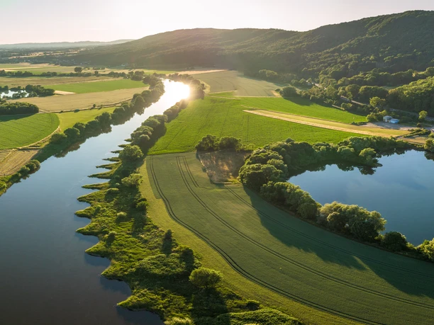 Porta Westfalica-Weserbogen-Teutoburger-Wald-Tourismus-D-Ketz-048.jpg Luftaufnahme eines malerischen Flusslaufs inmitten grüner Felder und bewaldeter Hügel bei Sonnenuntergang.